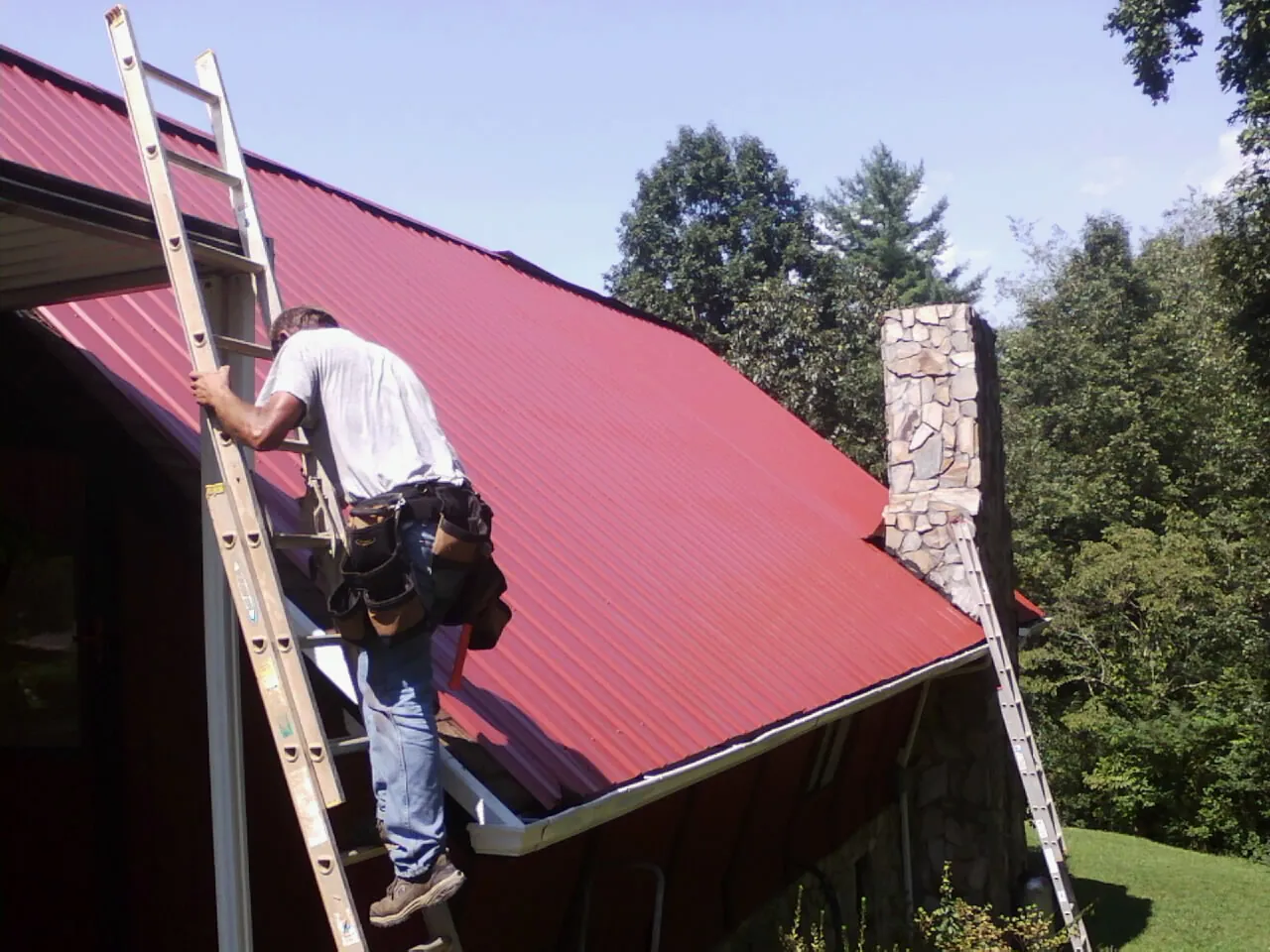 Roofer on a ladder beside a red metal roof and stone chimney — Soffit & Fascia Repair by Roofing Brothers in Vernal